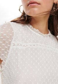 Blusa blanca texturizada con patrón de puntos en relieve, escote con volantes y delicado borde de encaje. Presenta mangas cortas y semi-transparentes.
