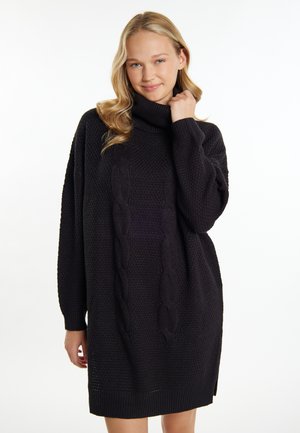 Jeune femme aux cheveux blonds portant une robe-pull col roulé tricotée en torsades noire, souriant doucement sur un fond blanc uni.