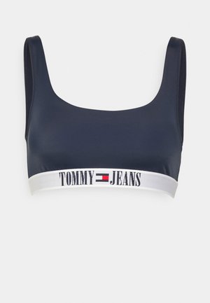 Jūras zilas krūšturis ar gludu tekstūru, kvadrātveida dekoltē un platu baltu joslu ar "TOMMY JEANS" logo melnā un sarkanā krāsā.