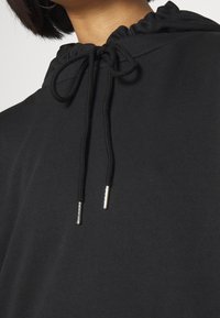 Sudadera negra con cuello fruncido y cordones. La tela es suave, con puntas metálicas en los cordones para un detalle adicional.