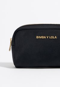 Juodas audinio maišelis su auksiniu logotipu "BIMBA Y LOLA", užtrauktuku, tiesių kampų forma, lygia tekstūra ir metaliniais akcentais.