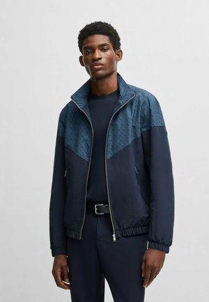 Marineblau und tealfarbene Zip-Jacke mit durchgehendem Reißverschluss, die durch einen gemusterten oberen Abschnitt und elastische Bündchen auffällt. Über einem dunklen Hemd getragen.