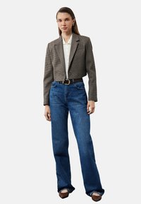Geknipt bruin houndstooth blazer, witte overhemd met knopen, hoge blauwe flare jeans en bruin platte schoenen. Model staat tegen een witte achtergrond.