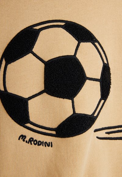 Μαύρη και μπεζ κεντημένη ποδοσφαιρική μπάλα πάνω σε μπεζ ύφασμα με τη λέξη "M.RODINI" γραμμένη κάτω από τη μπάλα.