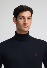 Maglione a collo alto blu navy in tessuto lavorato a maglia, con un piccolo emblema rosso sul lato sinistro del petto. Tessitura morbida con una silhouette aderente.