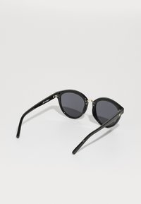 Ochelari de soare negri cu forma cat-eye, lentile închise, ramă din plastic lucios, accente aurii la balamale și brațe elegante, înguste.