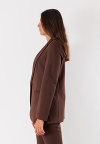 Donna con lunghi capelli castani in profilo, che indossa un blazer marrone e pantaloni abbinati su uno sfondo bianco semplice.