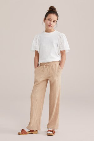 Teenagermädchen steht mit den Händen in den Taschen, trägt eine weiße Bluse mit Puffärmeln, beige strukturierte Hose, weiße Socken und braune Sandalen.