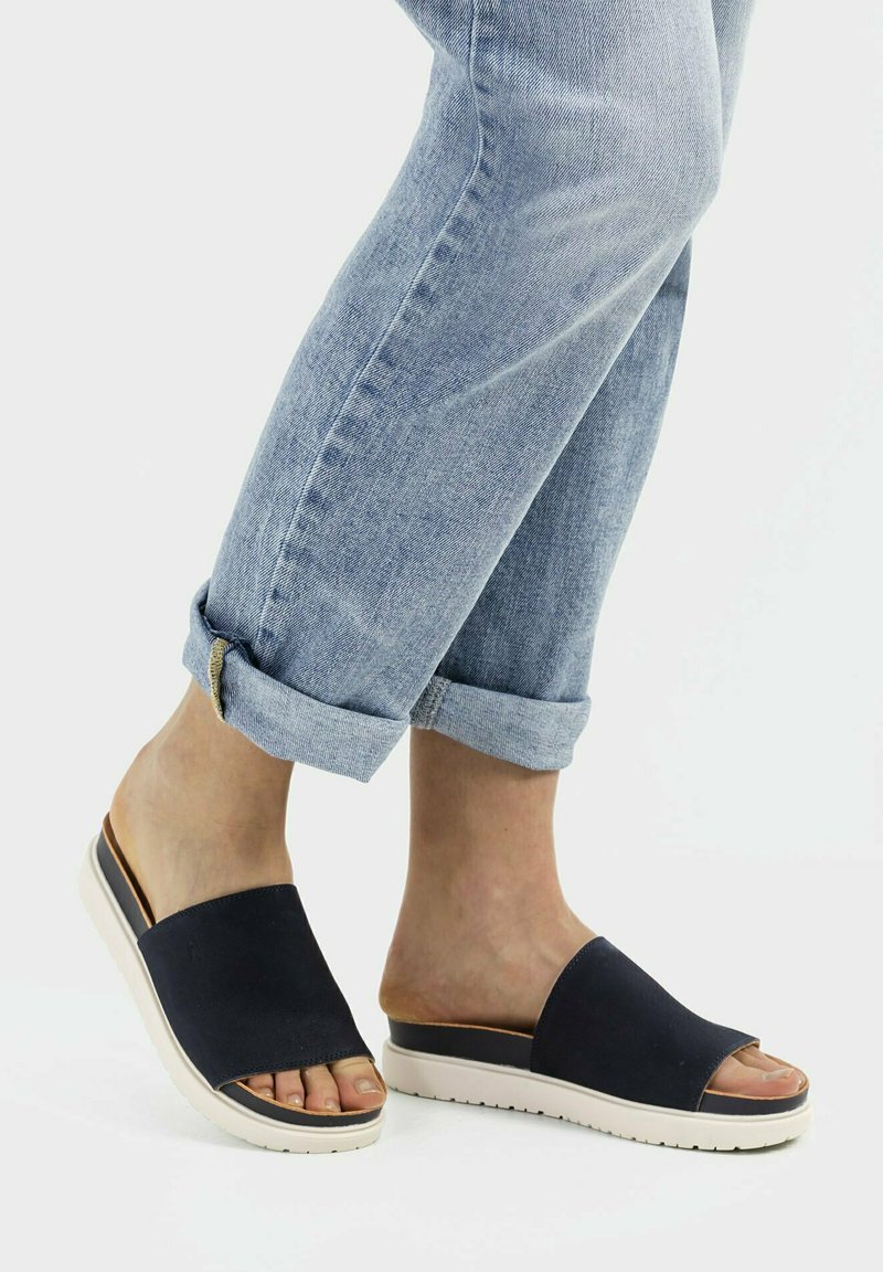 Navy slide sandals with a wide upper and open toe, featuring a textured sole and light-colored midsole, paired with rolled-up light blue jeans.