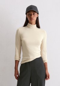Cremfarbener Rollkragenpullover mit langen Ärmeln, kombiniert mit dunkel gestreiften Hosen, ergänzt durch eine dunkelgraue Mütze. Glatte Textur und figurbetontes Design.