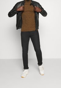 Persona che apre una giacca di pelle nera non zipata per rivelare un maglione marrone testurizzato, indossando pantaloni neri e sneakers bianche su uno sfondo semplice.
