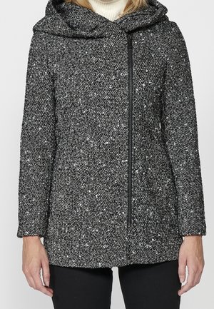 Vrouw draagt een zwart-wit gespikkelde wollen jas met een asymmetrische voorrits en een grote kraag over een witte coltrui.