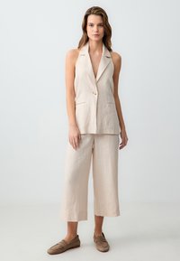 Beige linnen blazer met een diepe V-hals en enkele knoop, gecombineerd met bijpassende wijde cropped broek. Zachte textuur, minimalistisch ontwerp.