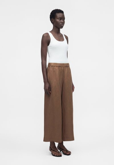 Γυναικείο μοντέλο φορώντας λευκό αμάνικο ριμπ τοπ, καφέ φαρδιά παντελόνια με ψηλή μέση και μαύρα σανδάλια με τακούνι, στέκεται σε λευκό φόντο.