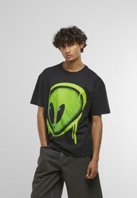 Sort bomulds t-shirt med et stort grønt aliengrafik og sorte detaljer. Løs pasform, korte ærmer og rund halsudskæring.