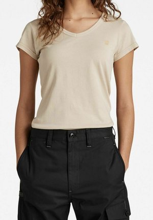 Kobieta w beżowej koszulce z krótkim rękawem w serek i czarnych spodniach cargo, z rękami w kieszeniach, na tle jednolitego białego tła.