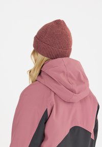 Vaaleanpunainen ja musta takki hupulla, yhdistettynä viininpunaisiin neulottuihin myssyihin. Teksturoitu myssy on pyöreä ylhäältä ja siinä on ribattu reuna.