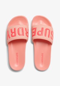 Sandales en caoutchouc roses avec une large lanière comportant le mot en relief "SUPERDRY" en corail contrasté. Semelle intérieure texturée pour le confort.