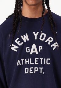 Sudadera azul marino con texto blanco: "DEPARTAMENTO DE ATLETIISMO DE NEW YORK GAP". Cuello redondo y mangas largas, hecha de una tela suave.