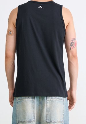 Person wearing a black sleeveless shirt with a small white logo on the upper back and light blue denim shorts, shown from behind.