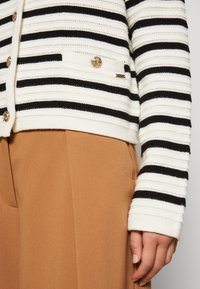 Gros plan d'une personne portant un cardigan en tricot crème et noir à rayures avec des boutons dorés et un pantalon couleur chameau.