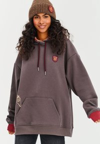 Hnedá mikina s predným vreckom, vínovými akcentmi a logom. Žebrované manžety a kapucňa so šnúrkou. Model má na sebe hnedú pletenú čiapku.
