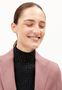 Hellrosa Blazer über einem schwarzen gerippten Rollkragenpullover mit weichem Textil. Die Form des Kragens und der maßgeschneiderte Schnitt sind in der Nahaufnahme gut sichtbar.