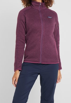 Vrouw die een paarse fleecevest met rits draagt en donkerblauwe broek, staand tegen een effen lichtgrijze achtergrond.