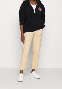 Sort fleece hættetrøje med halvdels lynlås og rød-hvidt logo. Kombineret med beige cargo bukser og hvide sneakers.