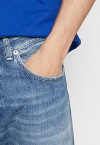 Jeans de mezclilla azul claro con un acabado desgastado, que presentan un bolsillo delantero y costuras amarillas. Una mano está apoyada en el bolsillo.