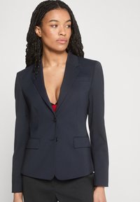 Femme aux longs cheveux tressés portant un blazer bleu marine ajusté sur un haut rouge, regardant légèrement vers sa gauche sur un fond uni.