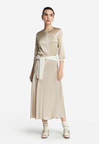 Beige satin midiklänning med långa ärmar, rund halsringning och knäppning, uppskuren midja med en vit tröja knuten. Ytterligare ljusa strumpor och skor.