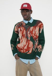 Πλεκτό πουλόβερ με πολύχρωμο σχέδιο αλόγου σε κόκκινο και μπεζ πάνω σε σκούρο πράσινο φόντο. Φορεμένο πάνω από ένα ανοιχτό γαλάζιο πουκάμισο με γιακά.