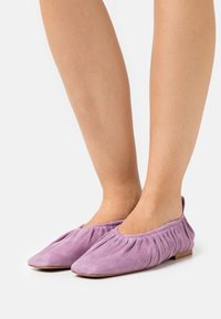 Scarpe flat in suede viola con un design elastico e arricciato sul tallone e una punta arrotondata. La suola è piatta e realizzata in materiale di colore chiaro.