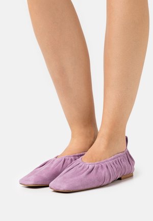 Zapatos planos de ante morado con un diseño elástico y recogido en el talón y una punta redondeada. La suela es plana y está hecha de un material de color claro.