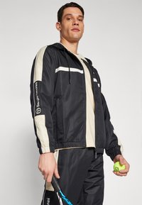 Chaqueta negra con rayas beige, material ligero, capucha, puños elásticos y logo en la manga. El modelo sostiene una raqueta de tenis y una pelota.