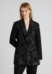 Blazer negro con un patrón floral de lentejuelas, solapa afilada, cierre de un solo botón y dos bolsillos frontales. La tela texturizada añade profundidad.