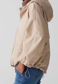 Giacca beige con cappuccio, chiusura a zip, polsini elastici e orlo con cordoncino. Realizzata in materiale liscio e resistente all'acqua; presenta tasche laterali.