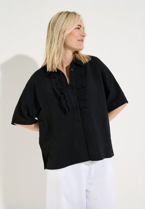 Blonde Frau, die nach rechts schaut, trägt eine lockere schwarze kurzärmelige Bluse mit Rüschen und weiße Hosen vor einem schlichten hellen Hintergrund.