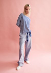 Long-sleeve striped top in blue and white, paired with light blue wide-leg pants featuring a tied waist. Model stands against a pink background.