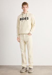 Jeune homme portant un sweat à capuche beige avec le texte "BOSS", un pantalon beige assorti et des baskets grises, se tenant devant un fond blanc uni.