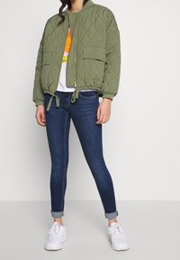 Chaqueta bomber acolchada de color verde oliva con cremallera frontal, puños elásticos, dos bolsillos frontales y dobladillo ajustable, combinada con jeans oscuros y zapatillas blancas.