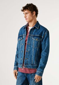 Chaqueta vaquera en azul medio con un corte clásico, que presenta dos bolsillos en el pecho, botones de metal y costuras en contraste. Se lleva sobre una camisa de cuadros roja.