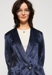 Sacou bleumarin dintr-un material neted și lucios, cu un design dublu și două butoane negre, purtat peste un top bej deschis.