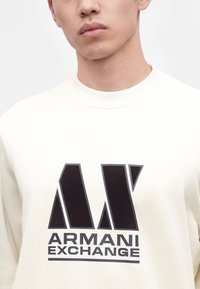 Homme portant un sweat-shirt blanc avec un grand logo noir Armani Exchange sur le devant, montré du cou au milieu du torse.