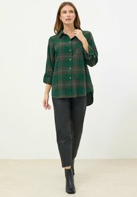 Camicia a quadri verde con maniche arrotolate, dotata di una tasca sul petto e orlo arrotondato. Abbinata a pantaloni neri e stivaletti.