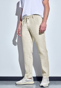 Pantalons en lin beige avec une taille élastique et des poches avant, associés à des baskets gris clair. Texture lisse et coupe décontractée.