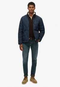 Man met een marineblauwe gewatteerde jas over een zwarte trui, blauwe jeans en tankleurige veterschoenen, staand met één hand in zijn zak.