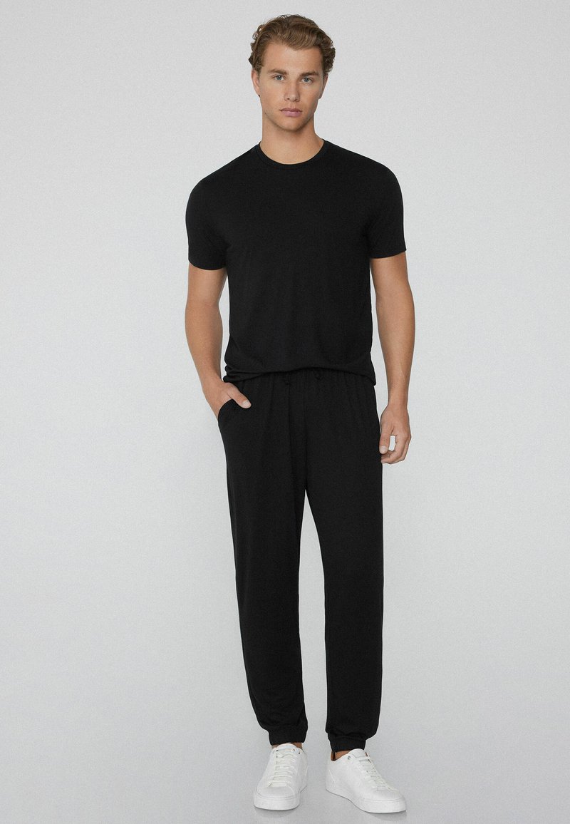 Schwarzes T-Shirt mit kurzen Ärmeln und schwarze Jogginghose mit Kordelzug. Glattes Material, normale Passform, elastische Bündchen an der Hose, kombiniert mit weißen Turnschuhen.