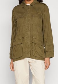 Oliwkowo-zielona kurtka użytkowa z przednimi kieszeniami na klapę, talią ze ściągaczem oraz beżowe spodnie cargo z bocznymi kieszeniami.